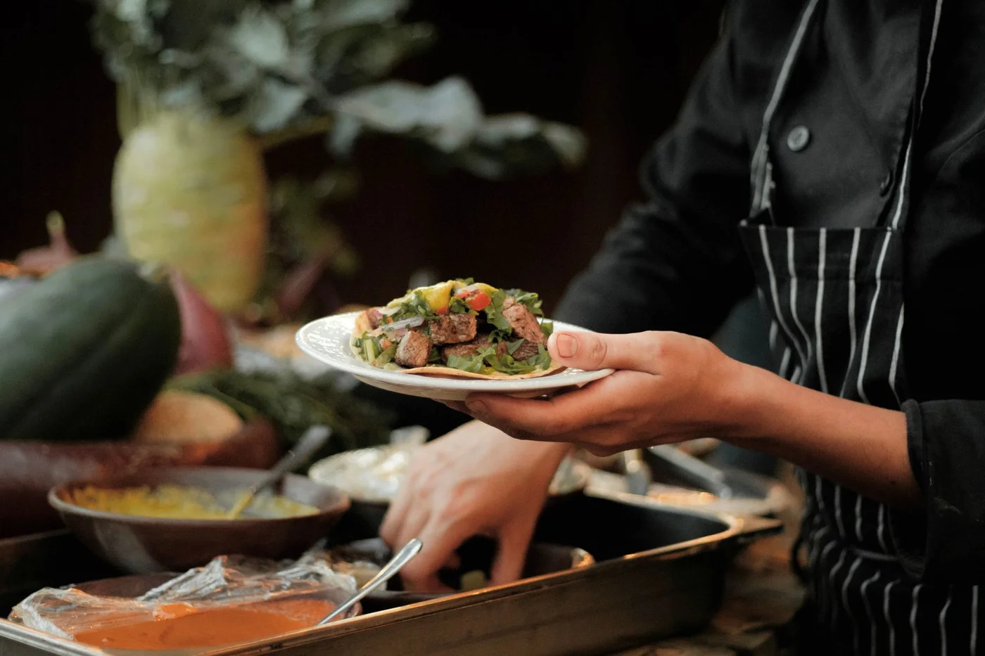 Private chef holding a garnished plate during an at-home service, seasonal vegetables in the background
